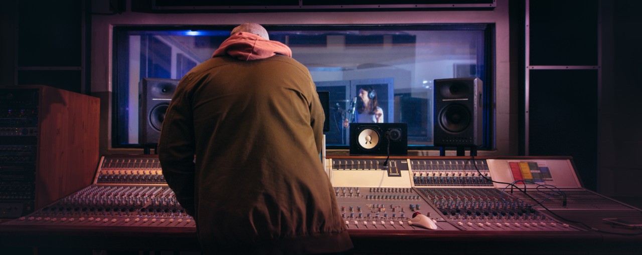 Sound engineer working at audio control panel with singer singing in recording room in background. Musicians producing music in professional recording studio.